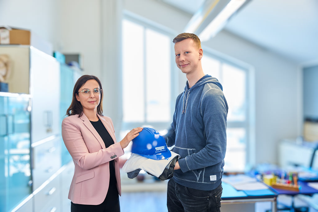 Bild von 2 Mitarbeitern, bei dem einer Person ein T.Shirt, eine Hose sowie ein blauer Helm mit dem Toepel Bau Logo überreicht wird